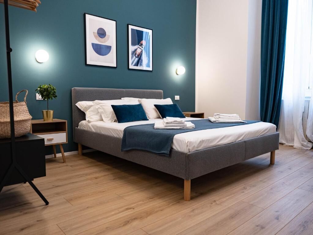 A modern bedroom with a grey bed, white linens, and blue pillows. There are two wall-mounted light fixtures and a framed picture above the bed.