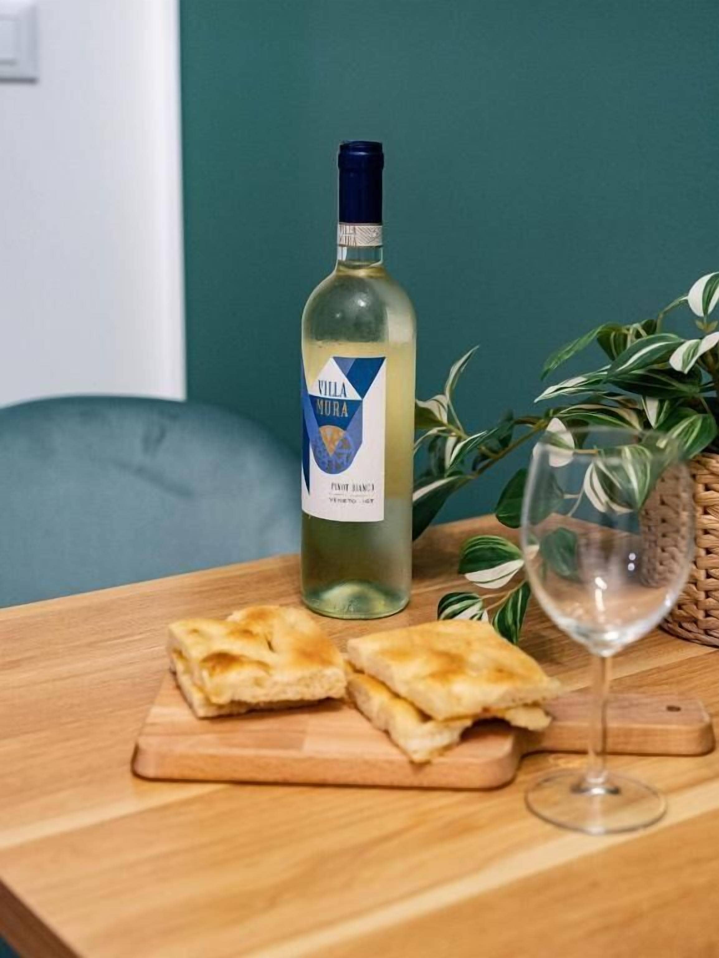 A bottle of Villa Mura Pinot Blanc, a glass of white wine, and two pieces of flatbread on a wooden table.
