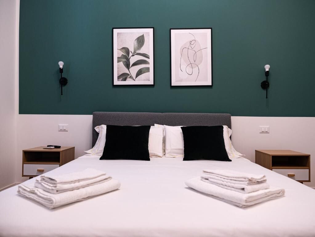 Una cama bien tendida con sábanas blancas y almohadas negras, dos cuadros enmarcados en la pared y mesitas de noche con lámparas.