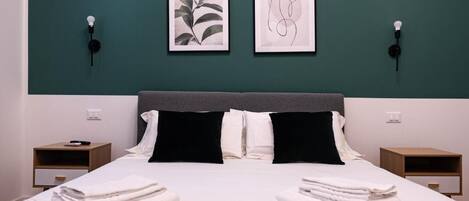 A neatly made bed with white linens and black pillows, two framed artworks on the wall, and bedside tables with lamps.