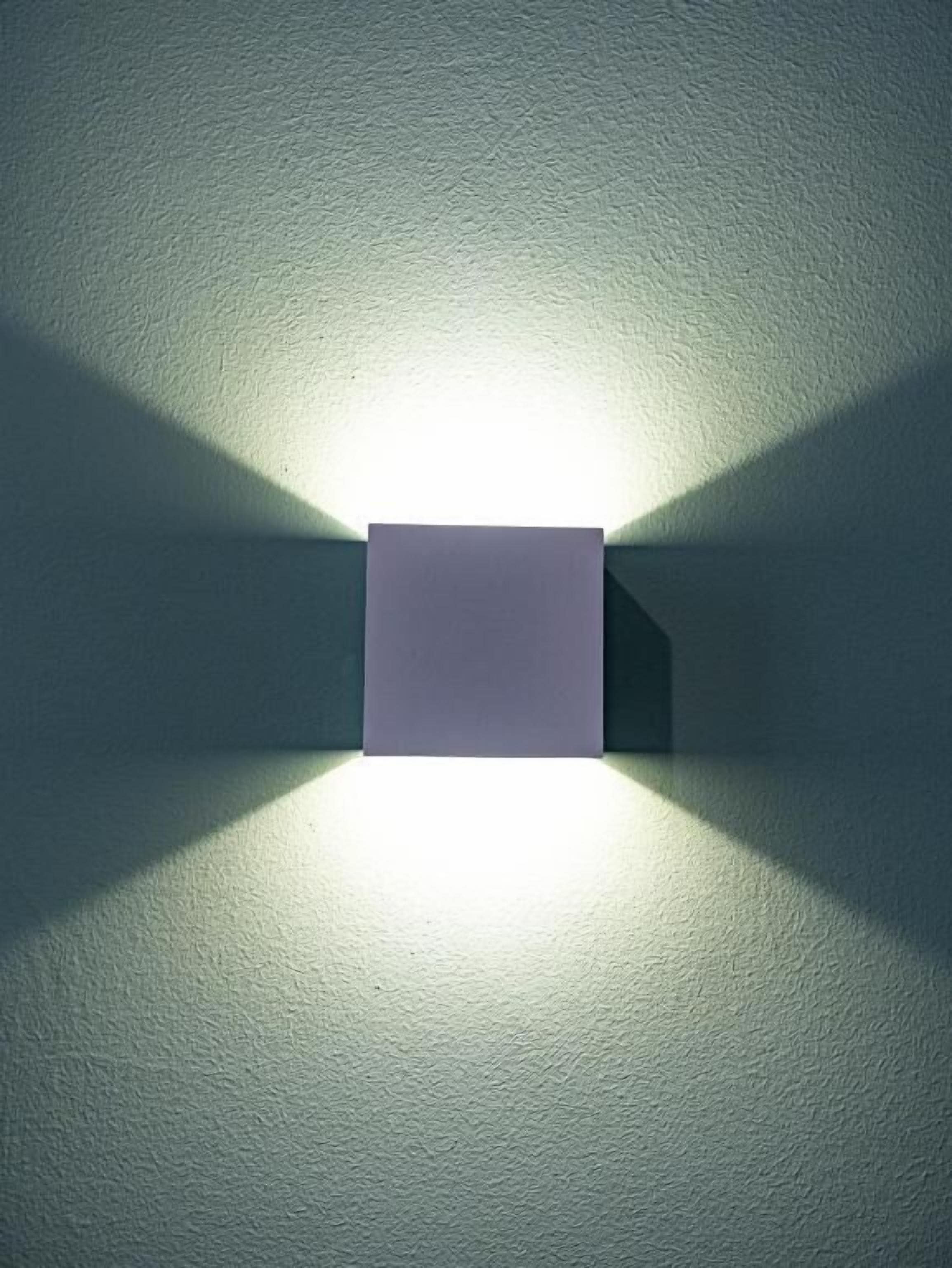 En väggmonterad lampa med en rektangulär skärm som ger ifrån sig ett sken och skapar en skugga på en vägg med tydlig struktur.