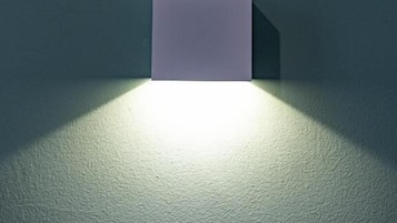 En väggmonterad lampa med en rektangulär skärm som ger ifrån sig ett sken och skapar en skugga på en vägg med tydlig struktur.