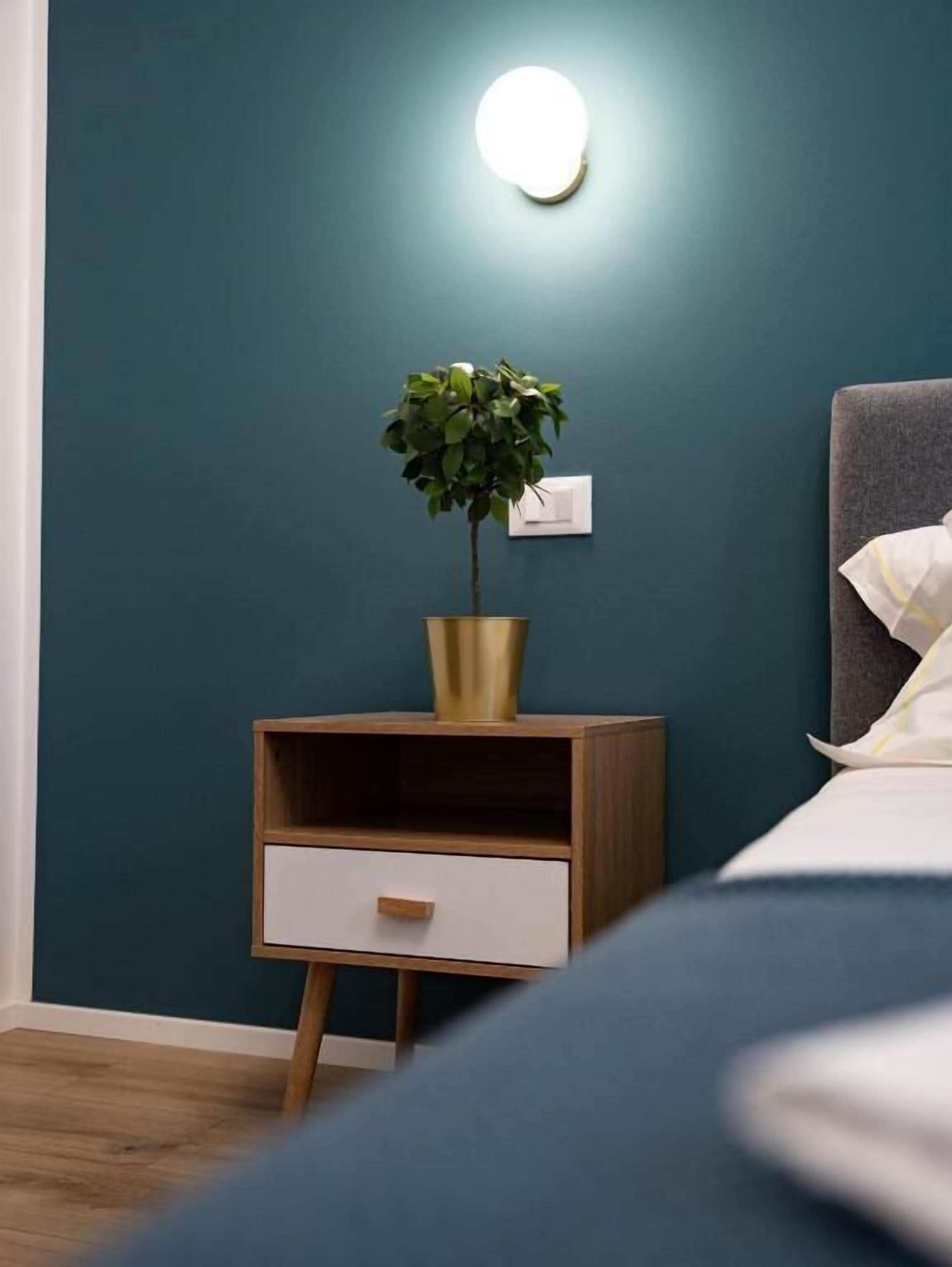 A bedroom with a bedside table, a nightstand, a potted plant, and a wall-mounted light.