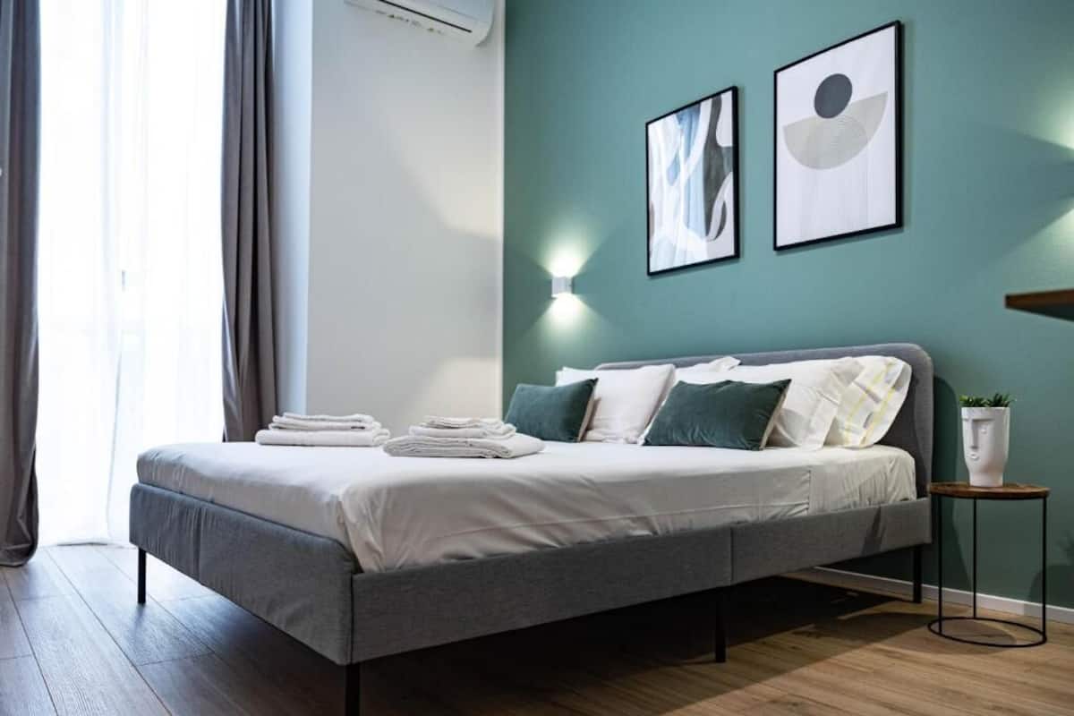 Ein modernes Schlafzimmer mit einem grauen Bett, weißer Bettwäsche, teal-farbenen Wänden und zwei gerahmten Kunstwerken.