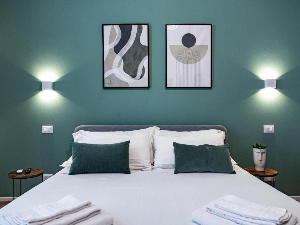 A neatly made bed with white and green bedding, two wall-mounted light fixtures, and two framed abstract artworks on a teal wall.