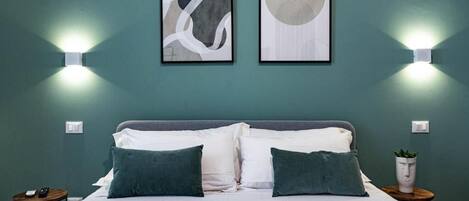 A neatly made bed with white and green bedding, two wall-mounted light fixtures, and two framed abstract artworks on a teal wall.