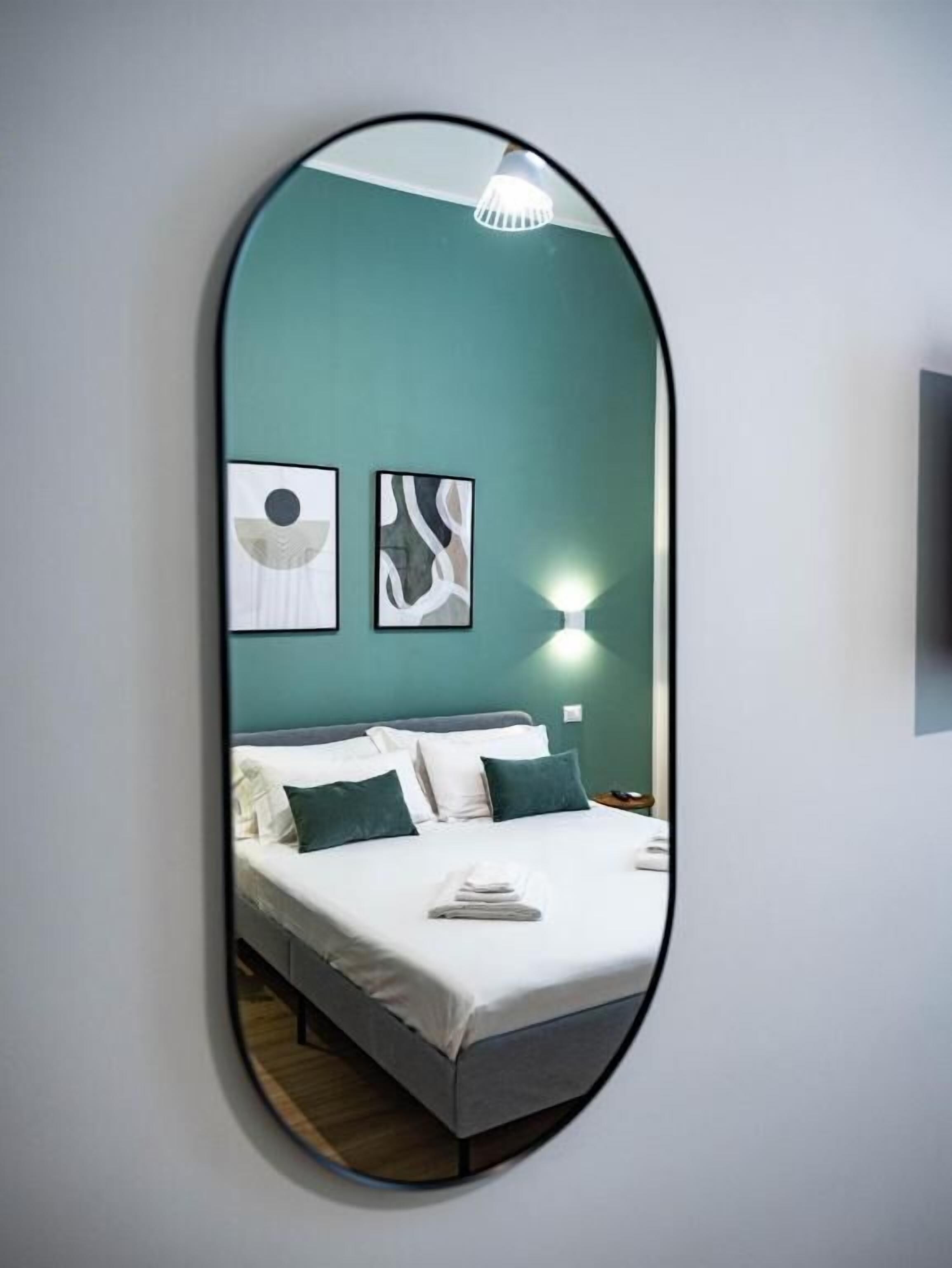 Een spiegel toont een bed met wit beddengoed en groene kussens, een wandlamp, en twee abstracte kunstwerken.