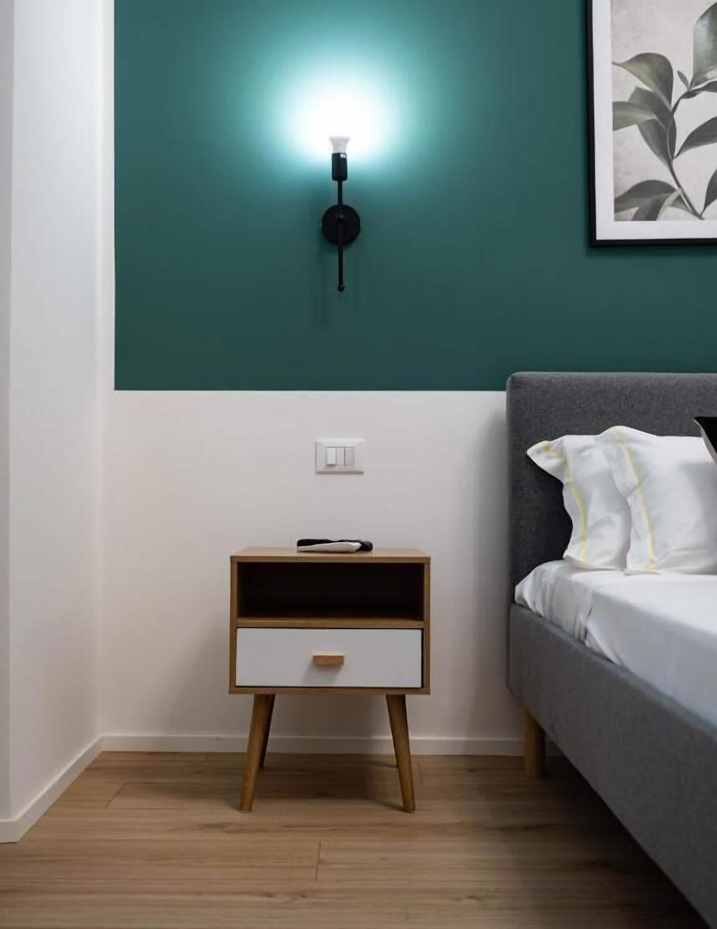 Un dormitorio con una pared de acento color turquesa, una mesita de noche con cajón, una cama gris con almohadas blancas y una lámpara fijada a la pared.