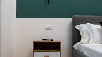 A bedroom with a teal accent wall, a bedside table with a drawer, a grey bed with white pillows, and a wall-mounted light.