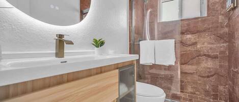 A modern bathroom with a white sink, wooden vanity, a glass shower enclosure, and a marble wall.