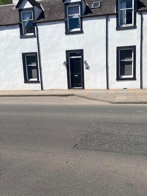 A white building with a black door and windows, a paved sidewalk, and a clear sky. - High Street House Sleeps 4 With Parking (Biggar)