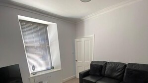 A living room with a black leather sofa, a television, a small table, and a window with blinds. - High Street House Sleeps 4 With Parking (Biggar)