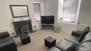 A modern living room with a black leather sofa, a flat-screen TV, a fireplace, and a large window with blinds. - High Street House Sleeps 4 With Parking (Biggar)
