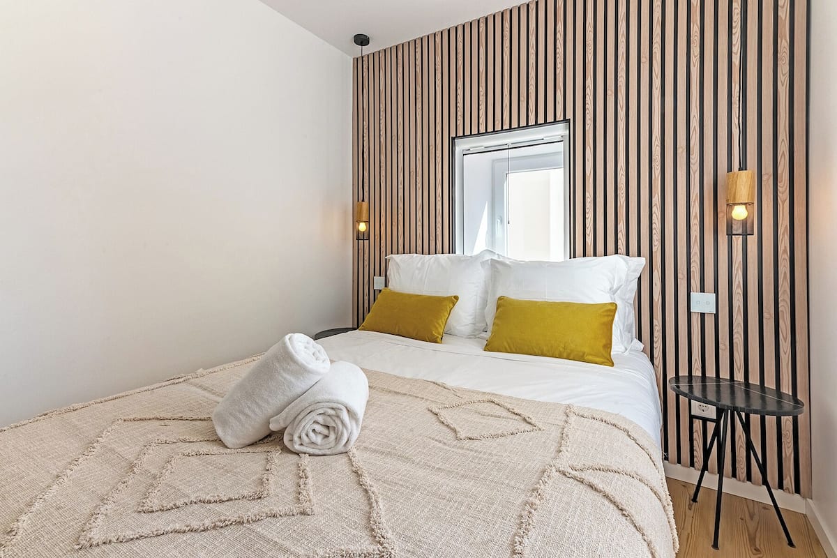 Quarto de hotel com uma cama, janela, luminária fixada na parede e uma pequena mesa.