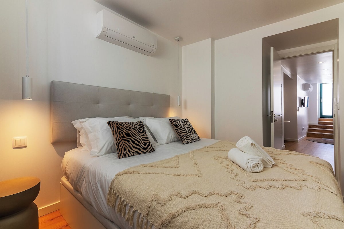 Quarto com cama, almofadas, cobertor, ar condicionado e porta que dá para outro cômodo.