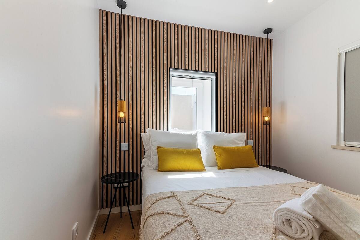 Quarto de hotel com uma cama, uma mesinha, duas luzes fixadas na parede e uma janela.