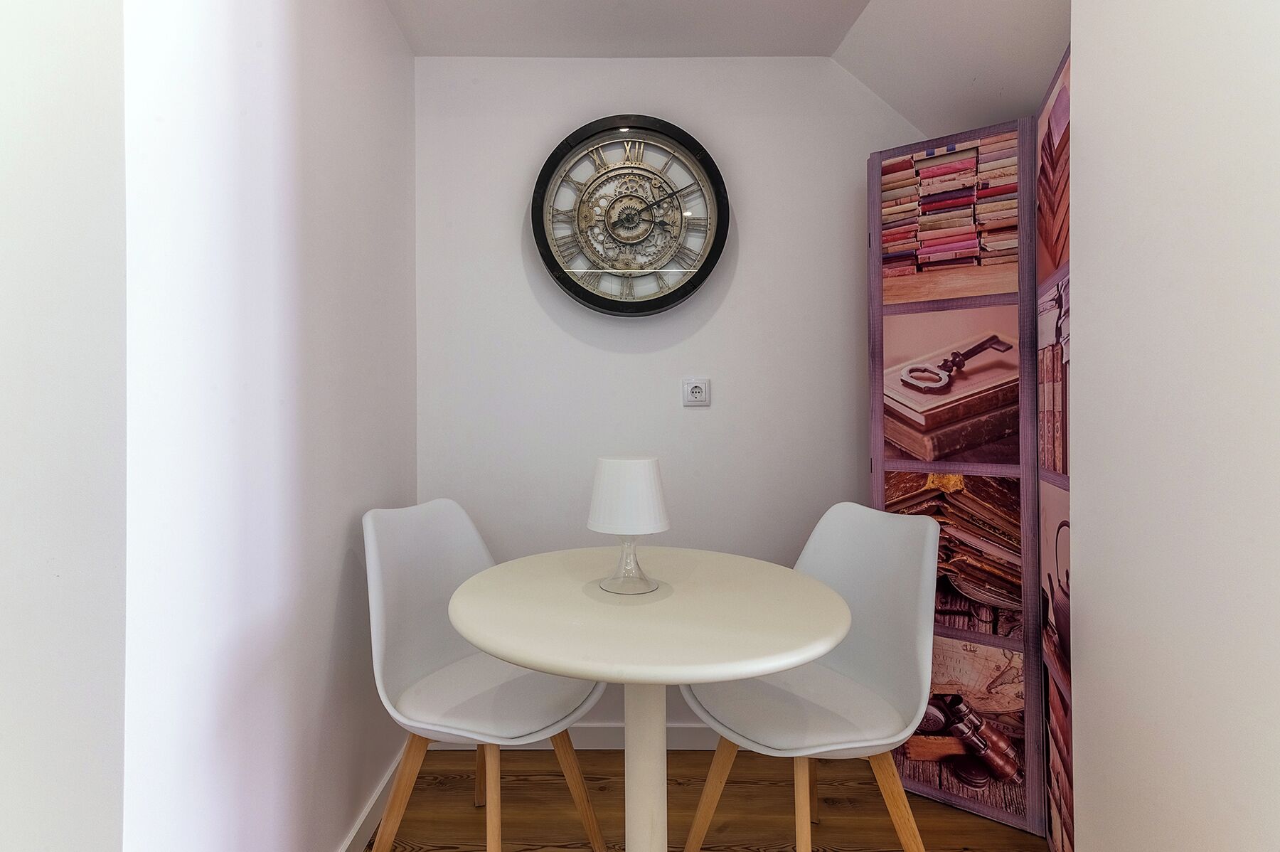 A small room with a round table, two chairs, a wall clock, and a bookshelf with books and decorative items.
