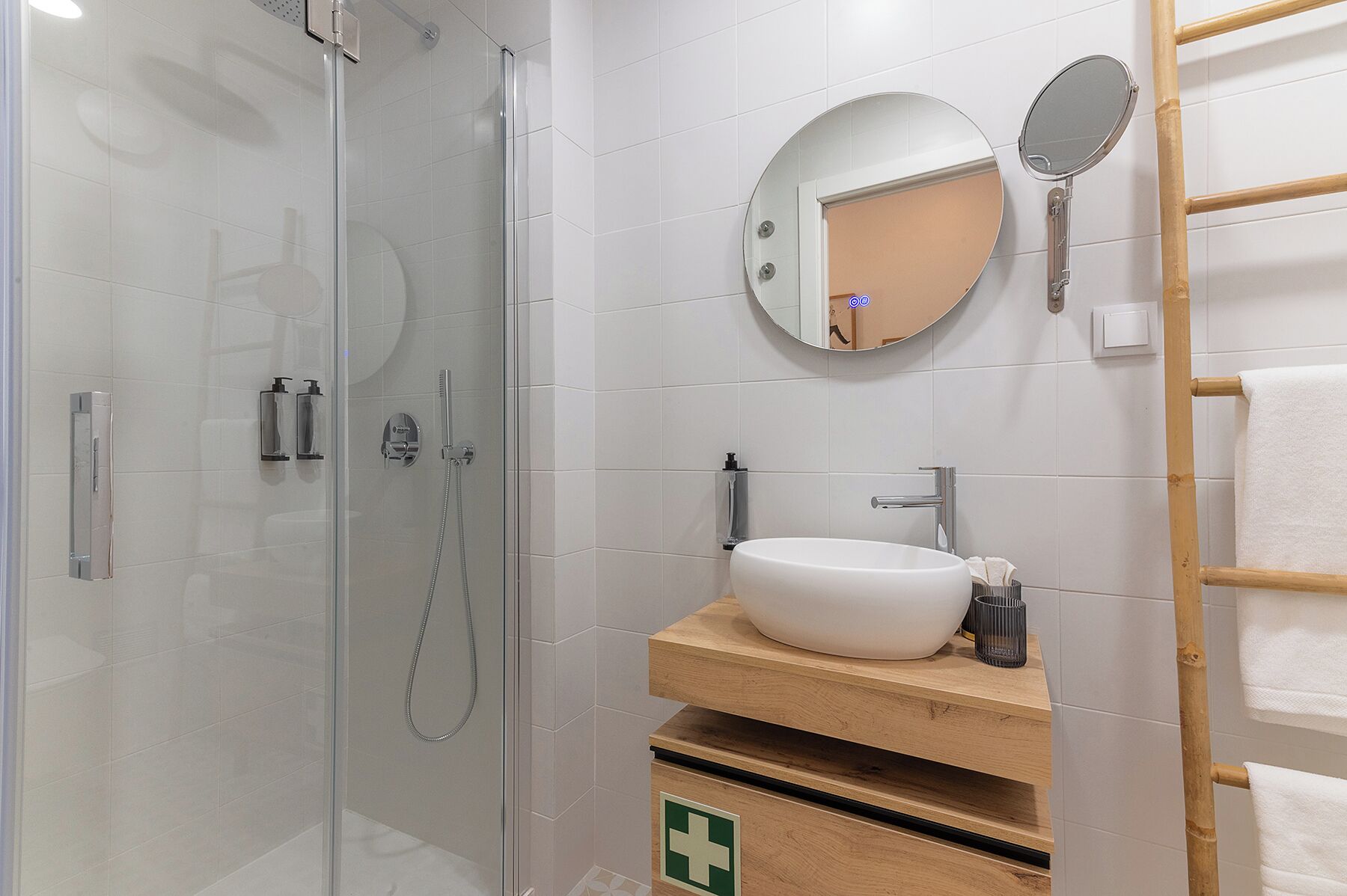 Une salle de bain moderne avec une douche à l’italienne en verre, un lavabo blanc doté d’un plan de toilette en bois, un miroir rond et un porte-serviettes.