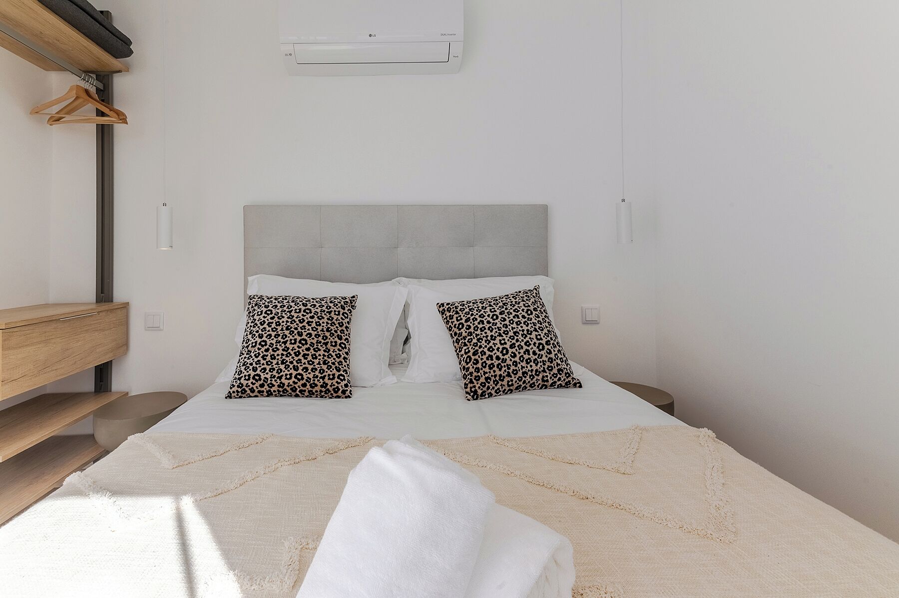 A bedroom with a bed, two pillows, a bedside table, a hanging light, and a wall-mounted air conditioner.