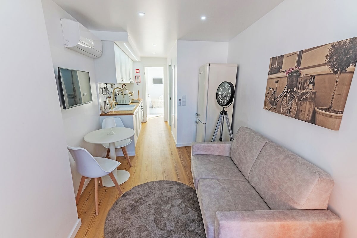 Uma sala de estar moderna com um sofá, um tapete redondo, uma área de jantar com mesa e cadeiras, e uma cozinha visível ao fundo.