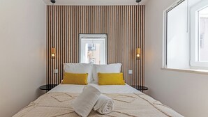 A neatly made bed with a rolled towel, two bedside tables, a window, and a view of the outside. - White Angels by Portas do Sol Apartments (Lisbon)