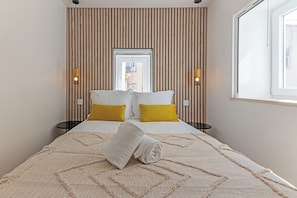 Ein ordentlich bezogenes Bett mit einem zusammengerollten Handtuch, zwei Nachttische, ein Fenster und Blick ins Freie.