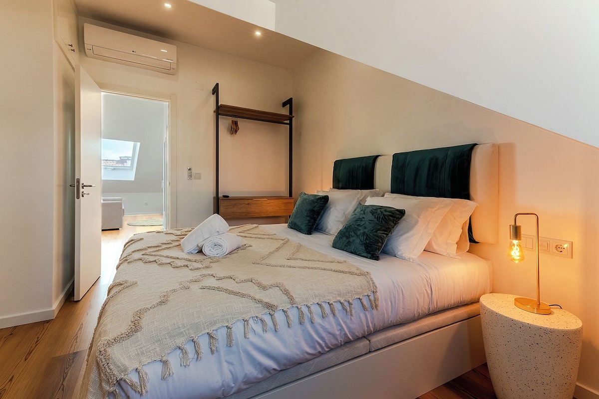 Quarto com uma cama, mesa de cabeceira, luminária pendente e porta que dá para outro cômodo.