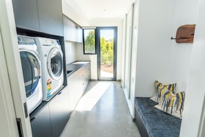 A modern laundry room with built-in washing machines, a bench with pillows, and a view through a door to the outdoors. - MAGNIFICENT AND OPULENT 5 BEDROOM OASIS BOASTING SWEEPING VALLEY VIEWS (Yallingup)