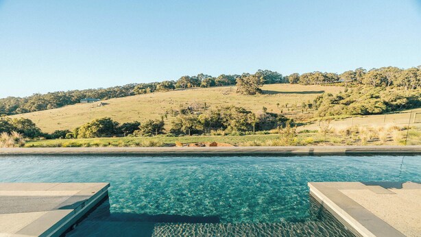 Una piscina con acqua cristallina, circondata da una pavimentazione in cemento, con vista su un paesaggio collinare con alberi e una recinzione sullo sfondo.
