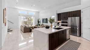 Cozinha moderna com ilha central, eletrodomésticos de aço inoxidável e área de refeições com sofá e mesa.
