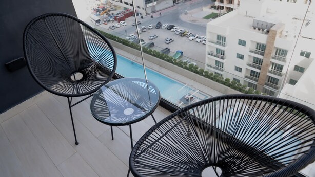 Varanda com três cadeiras de arame preto e uma mesa pequena, com vista para uma rua e edifícios da cidade.