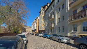 Une rue pavée bordée d’immeubles à plusieurs étages, de voitures garées et d’un arbre.