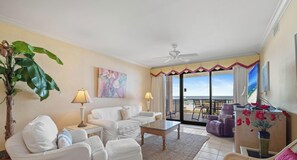 Salón con sofá, sillones, una mesa de centro, una planta y un cuadro. Hay un balcón con vistas al mar.