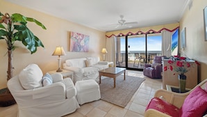 A living room with a white sofa set, a coffee table, a large potted plant, and a view of the ocean through the balcony doors. - Romar House 203A by Liquid Life (Orange Beach)