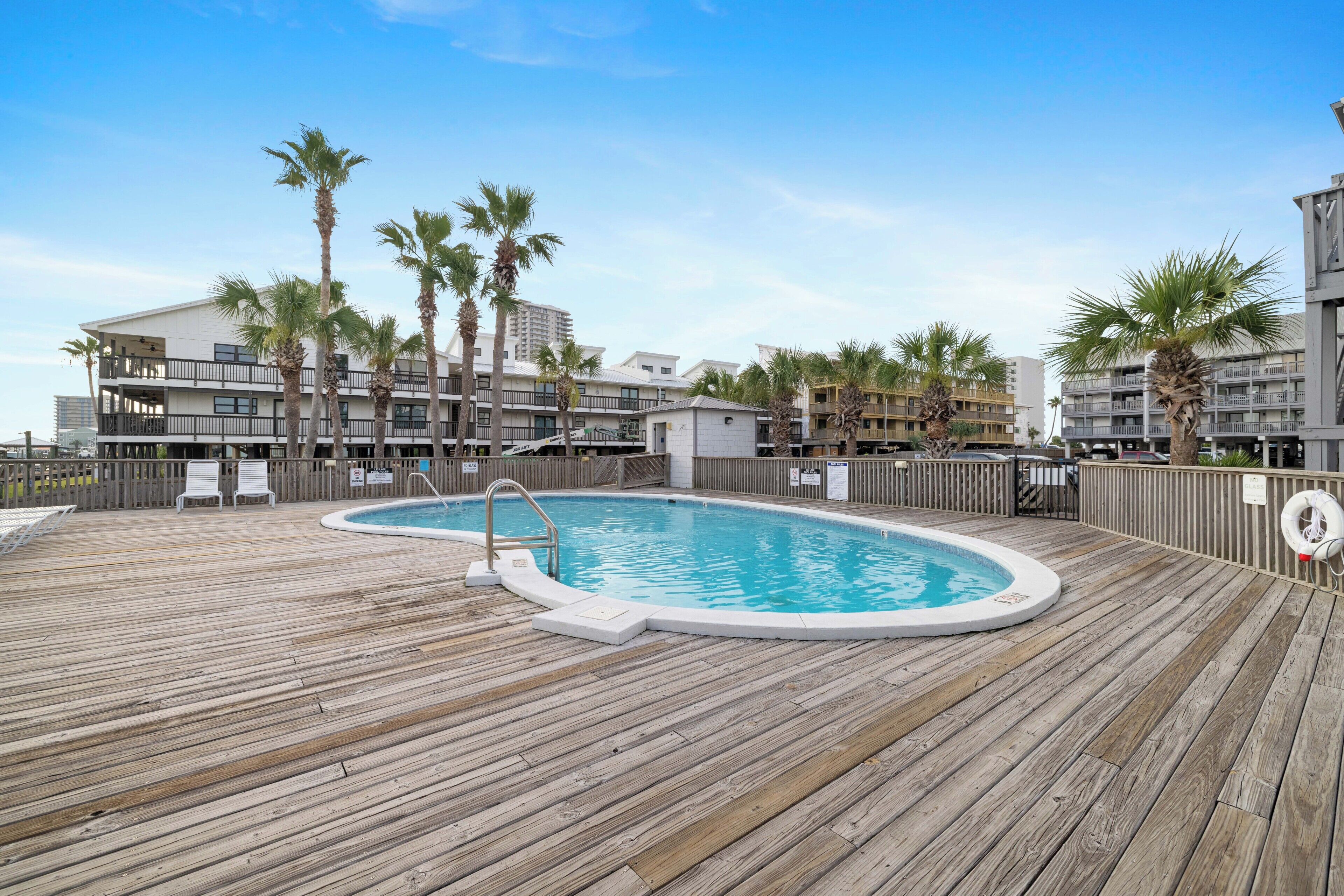 Eine Holzterrasse mit Swimmingpool, Palmen und einem mehrstöckigen Gebäude im Hintergrund.
