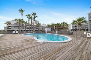 A wooden deck with a swimming pool, palm trees, and a multi-story building in the background. - Lei Lani Mini Tower 315N by Liquid Life (Orange Beach)