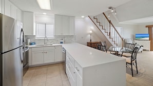 A modern kitchen with a central island, a dining area with chairs, a staircase, and a TV in the living room. - Lei Lani Mini Tower 315N by Liquid Life (Orange Beach)