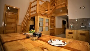 A wooden dining table with a plate, a glass, and a bowl. There is a wooden staircase and a kitchen area in the background. - Mountainshack Riverside Stay & Cafe (Kullu)