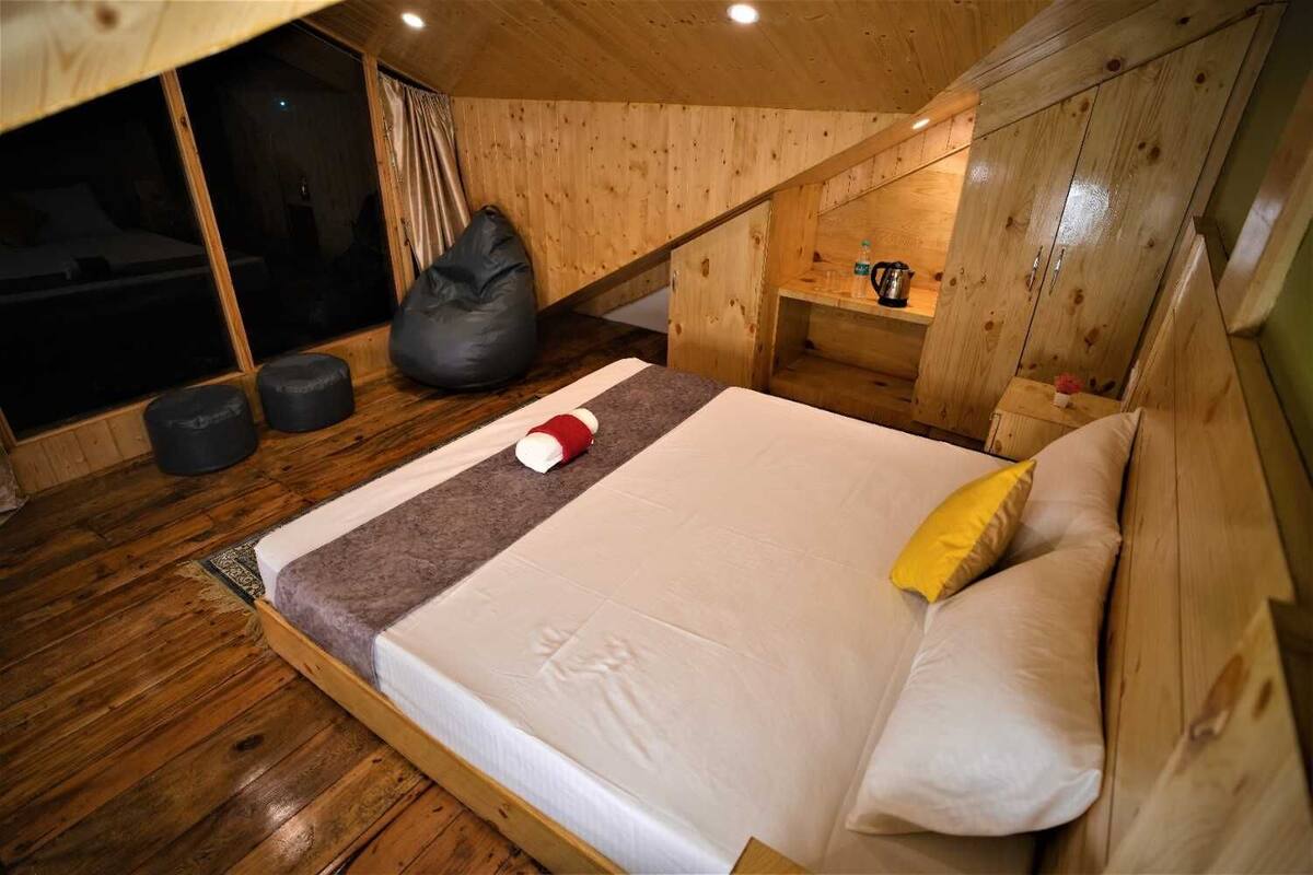 Un dormitori de cabana de fusta amb un llit, almoines, una taula petita amb un brollador i una finestra amb cortines.