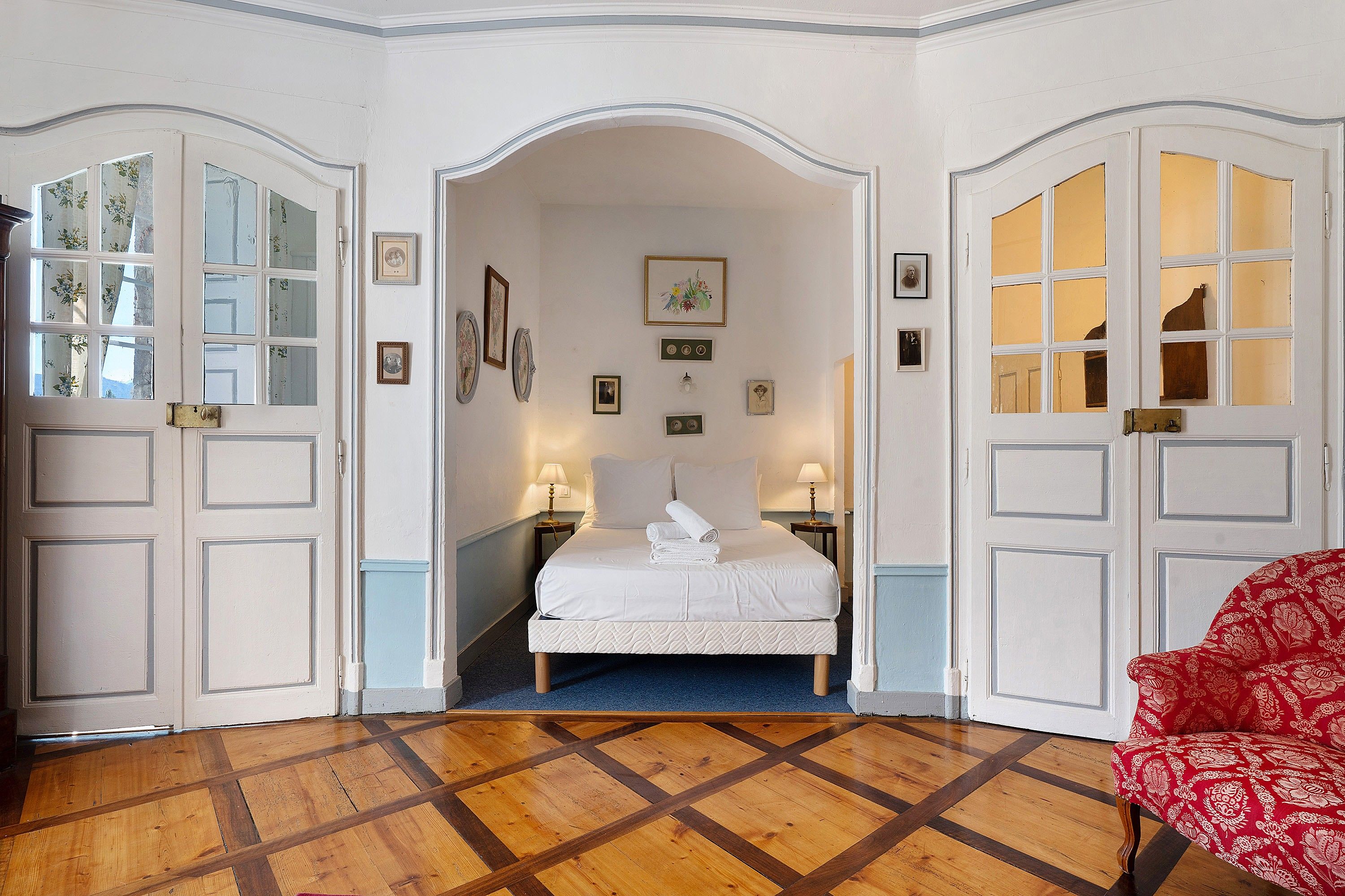 A hotel room with a bed, two bedside tables, a red patterned chair, and a wooden parquet floor.