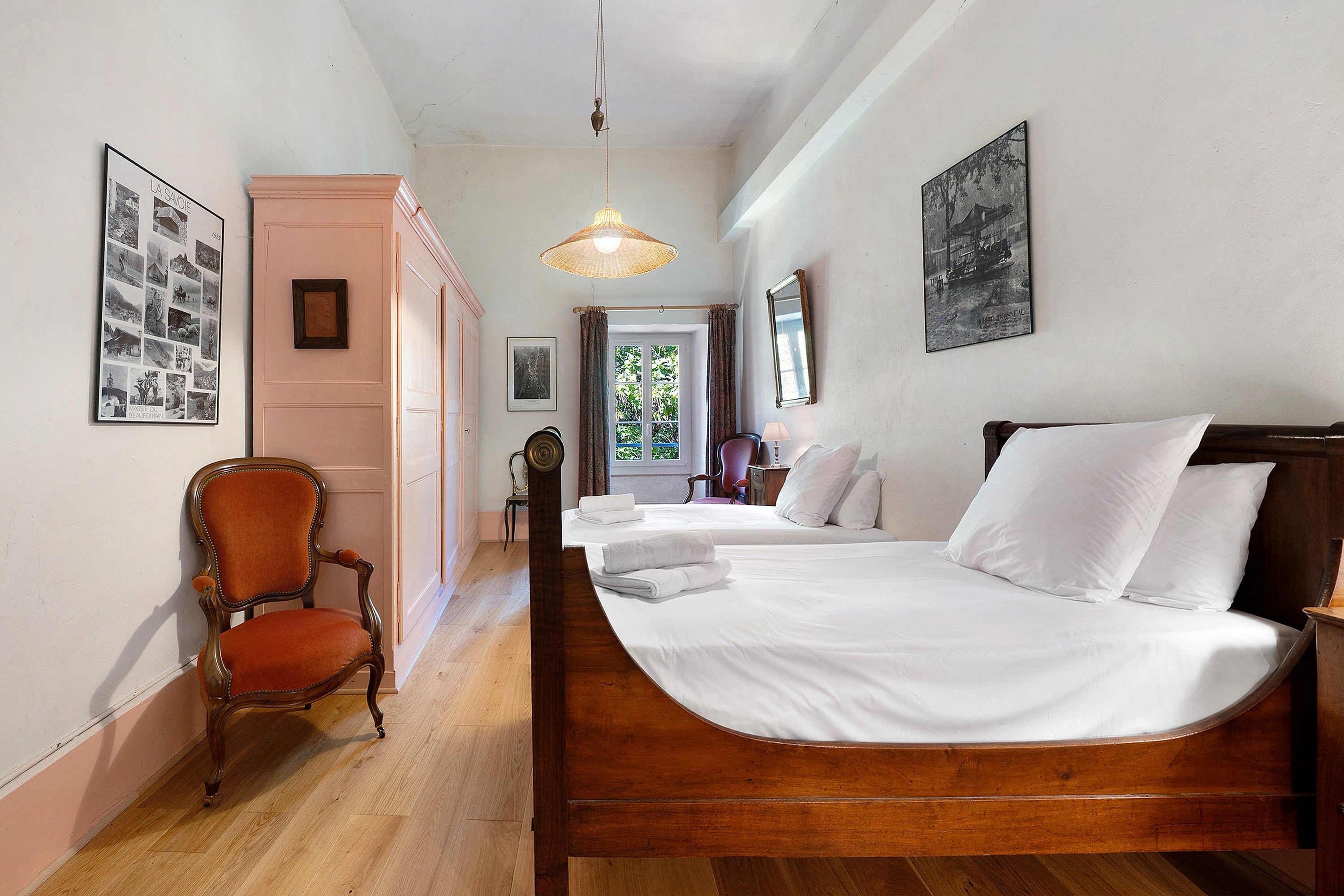 A hotel room with a wooden bed, a chair, a hanging light fixture, and framed pictures on the walls.