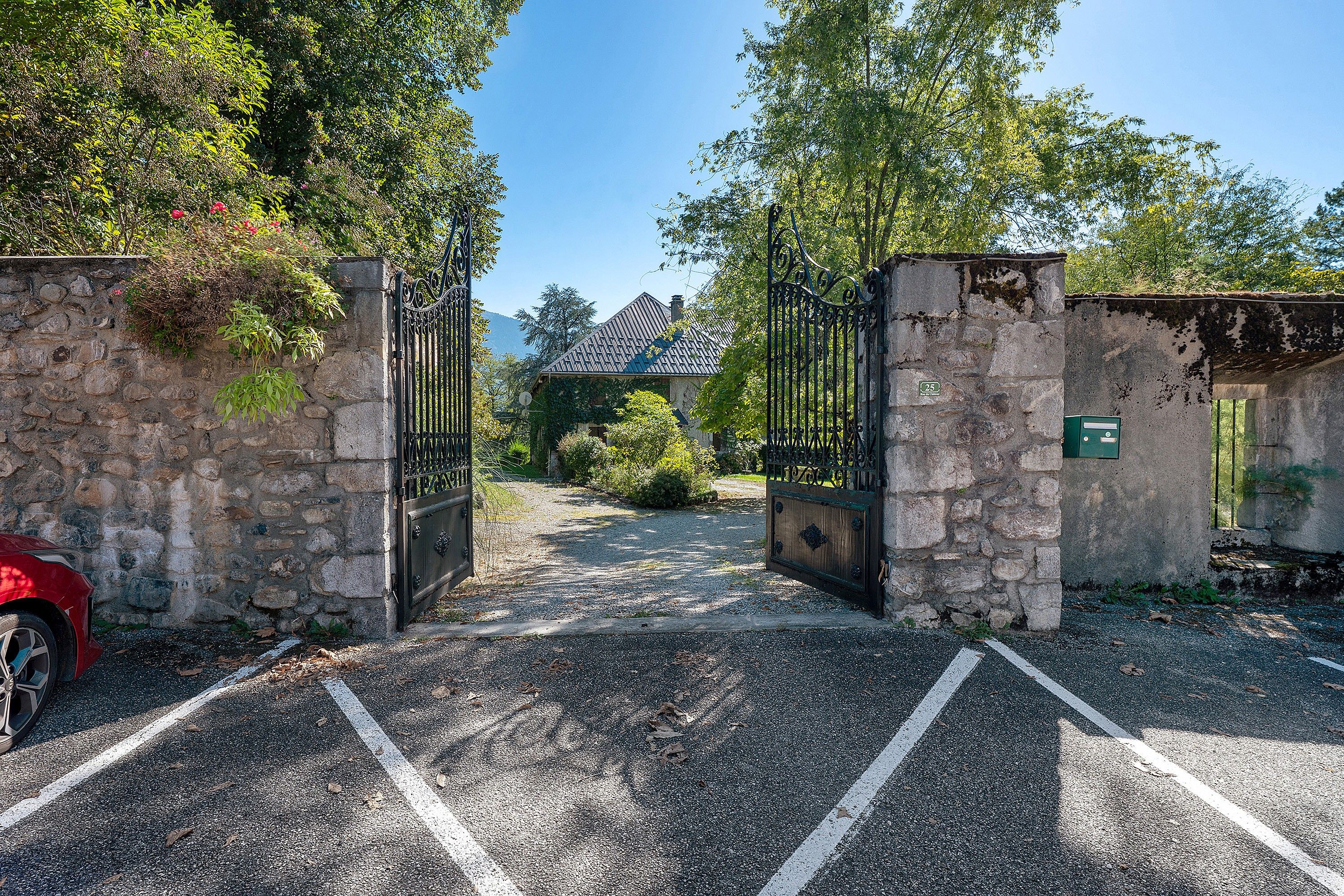 Une entrée fermée par un portail, un mur de pierre, une allée gravillonnée et, au fond, un bâtiment avec un toit recouvert de tuiles.
