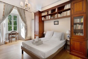A neatly arranged bedroom with a large bed, a wooden bookshelf, a window with curtains, and a small table with a chair. - Château with Private Park – Gîte du Tilleul – Exceptional Setting (Tournon)