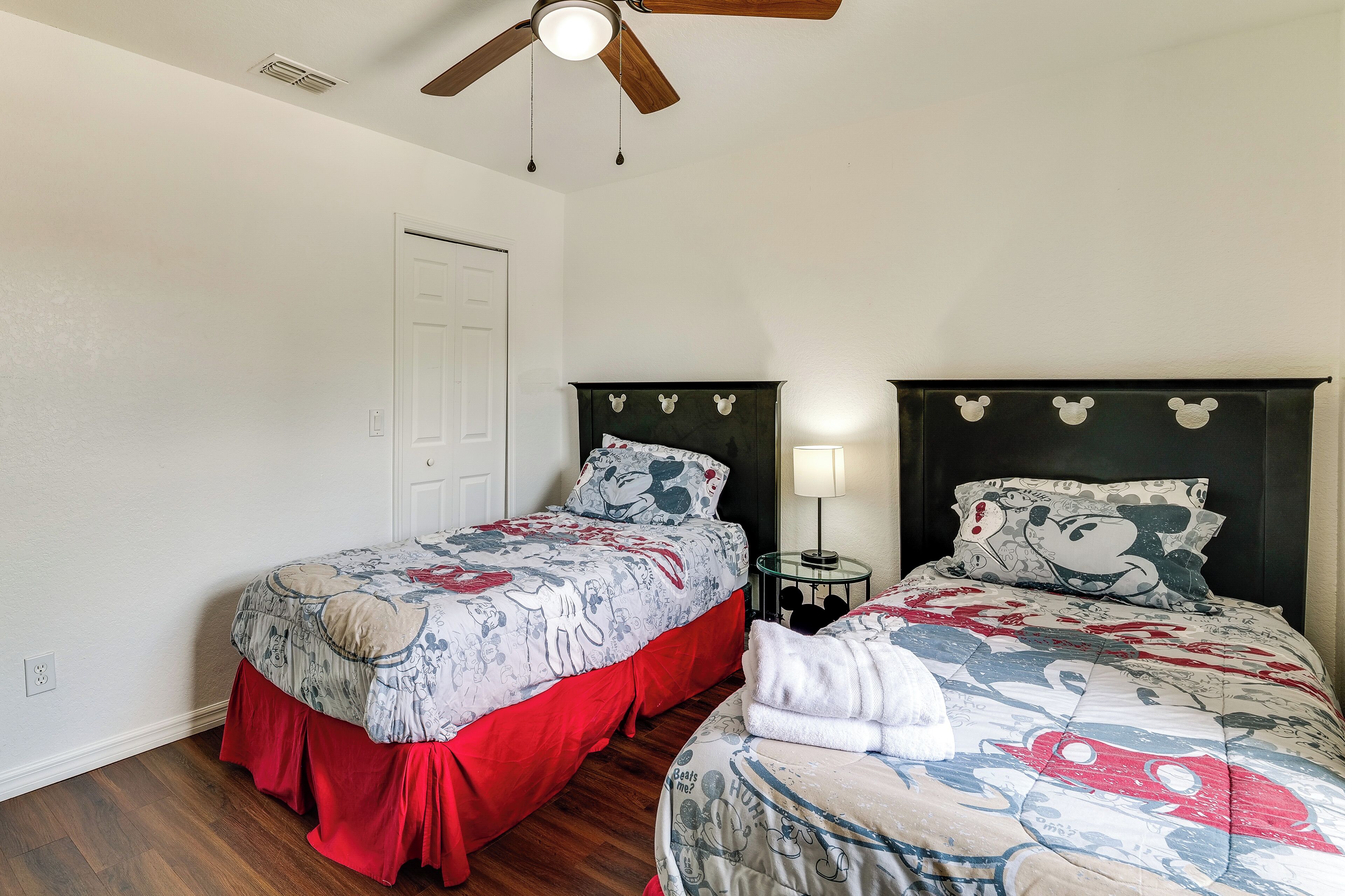 A bedroom with two beds, a ceiling fan, a nightstand with a lamp, and a door.