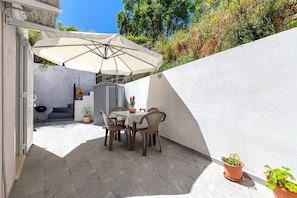 En udendørs terrasse med et bord, stole, en stor parasol og krukker med planter.