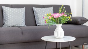 Ein modernes Wohnzimmer mit einem grauen Sofa, zwei gemusterten Kissen, einer weißen Vase mit Blumen und einem kleinen runden Tisch.