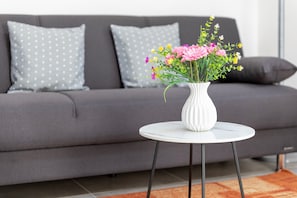 A modern living room with a grey sofa, two patterned pillows, a white vase with flowers, and a small round table. - Apartment 'Casa Angela' with Private Garden, Balcony and Air Conditioning (Nulvi)