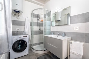 A modern bathroom with a glass-enclosed shower, a washing machine, and a sink with a mirror. - Apartment 'Casa Angela' with Private Garden, Balcony and Air Conditioning (Nulvi)