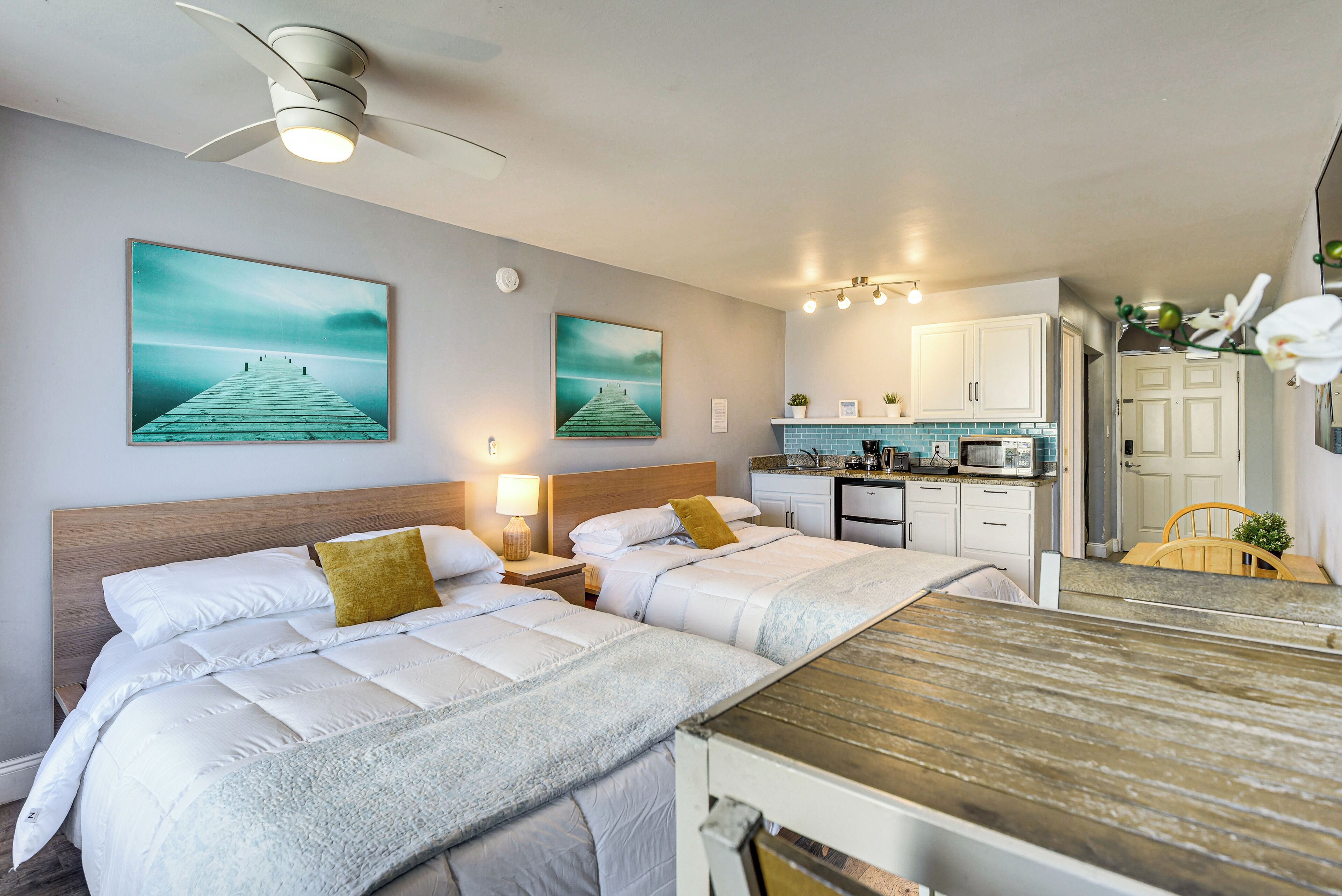 Ein Hotelzimmer mit zwei Betten, einem Holztisch, einer Küchenzeile und Bildern an den Wänden.
