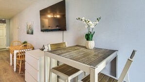 Un coin repas avec une table en bois, des chaises blanches, un téléviseur à écran plat fixé au mur et un vase avec des fleurs blanches.